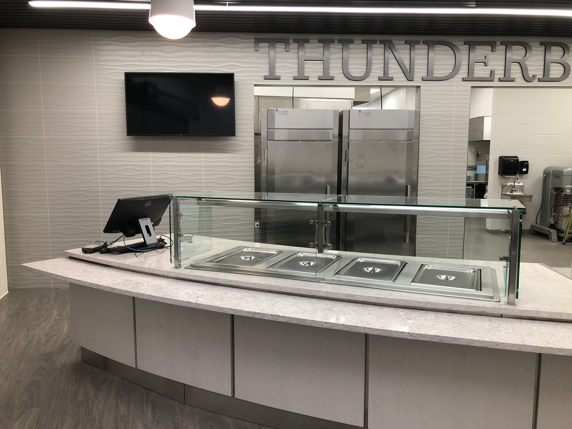 A food service counter with a stainless steel sneeze guard and a television on a light gray tiled wall.