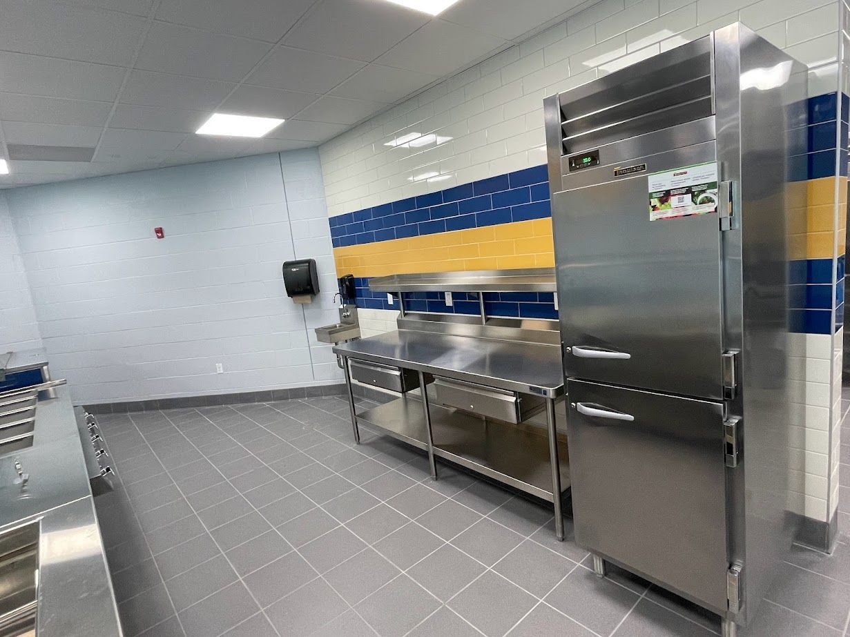 Commercial kitchen with a stainless steel refrigerator, metal prep tables, and blue, yellow, and white tiled walls.