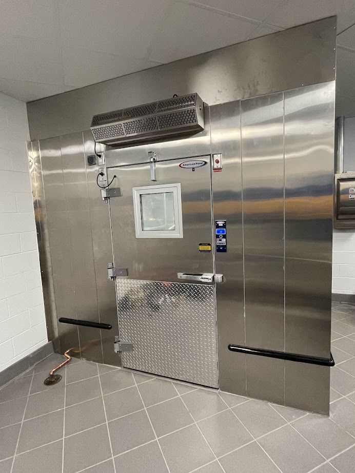 A commercial stainless steel walk-in cooler door with a small window, diamond-plate kick plate, and an overhead air curtain.