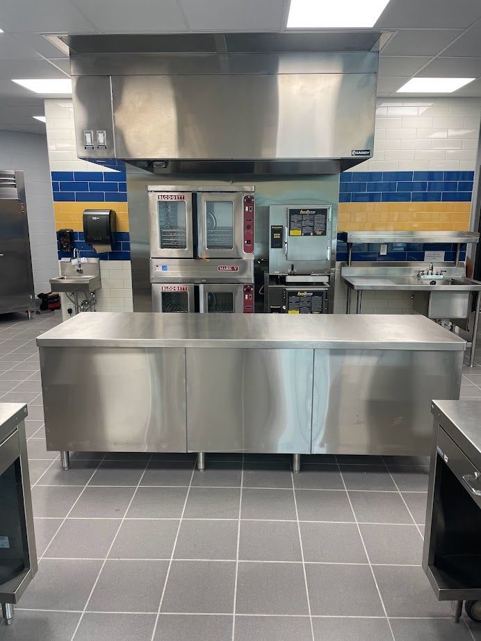 A commercial kitchen featuring a central stainless steel island, double ovens beneath a large hood, and tiled walls.