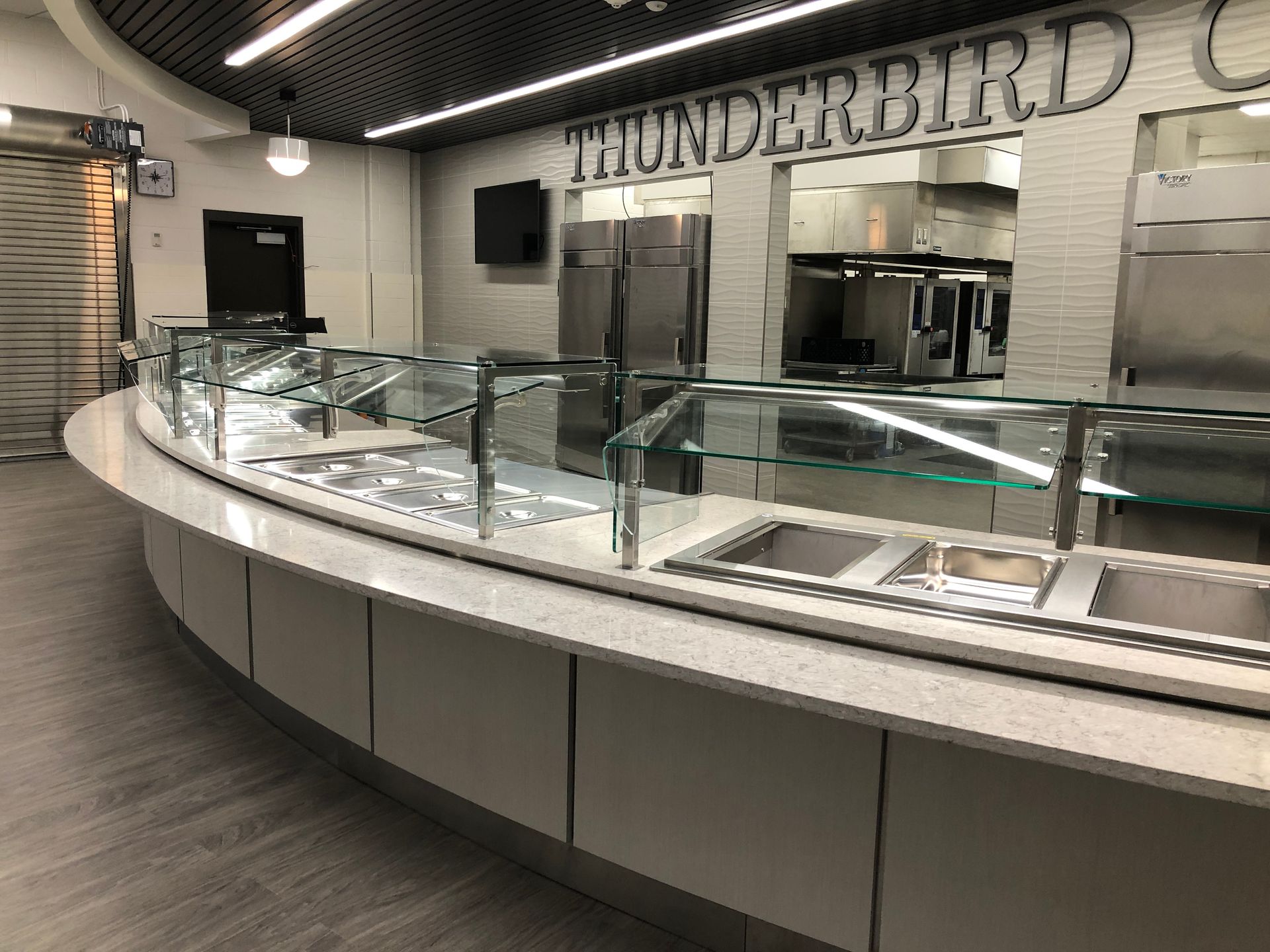 A curved food service station counter with a glass sneeze guard, metal serving trays, and a 