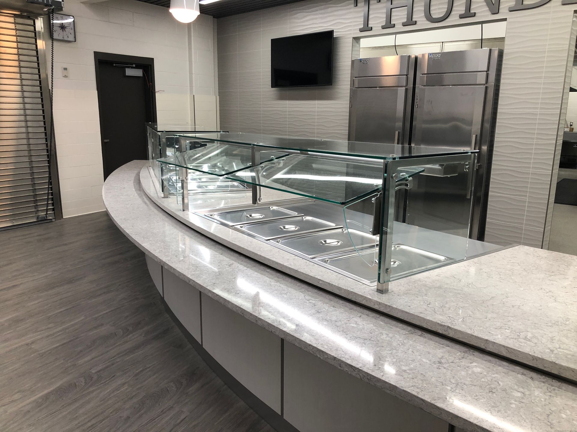A curved white stone counter with a glass sneeze guard and stainless steel food warmers in a modern cafeteria.