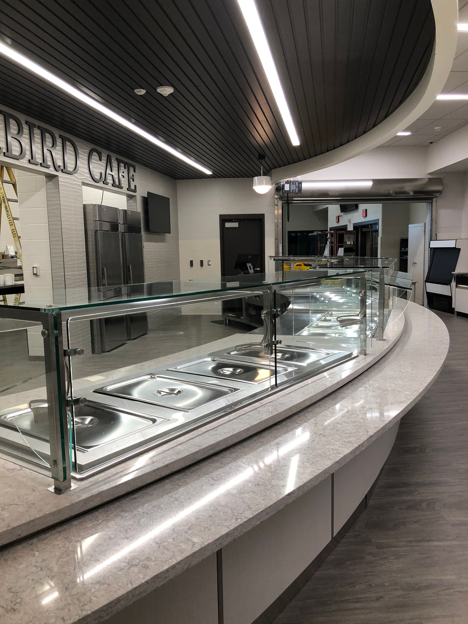 A curved cafeteria service counter with stainless steel food warmers and glass guards beneath a modern black ceiling.
