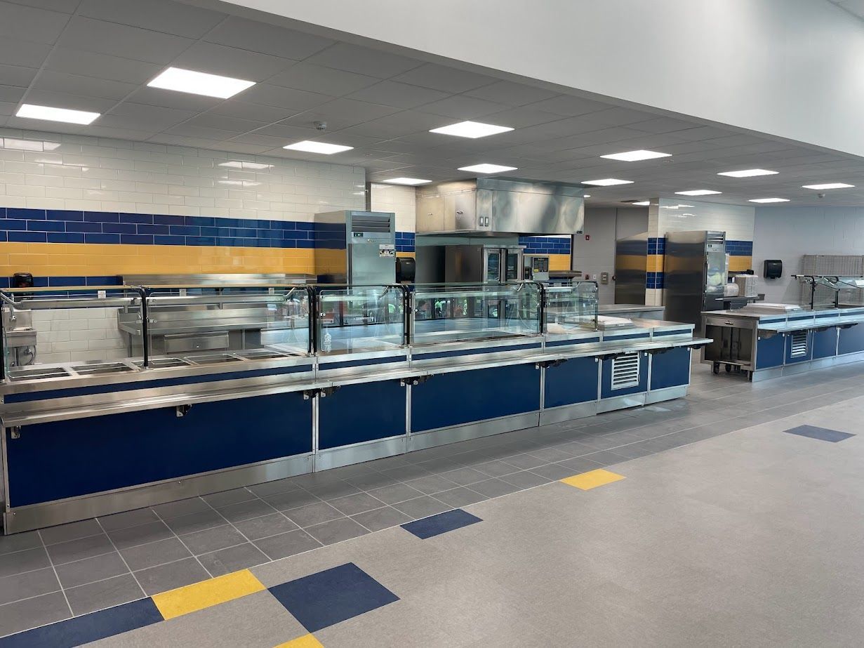 A modern school cafeteria serving line with blue and yellow wall tiles, stainless steel counters, and checkered flooring.