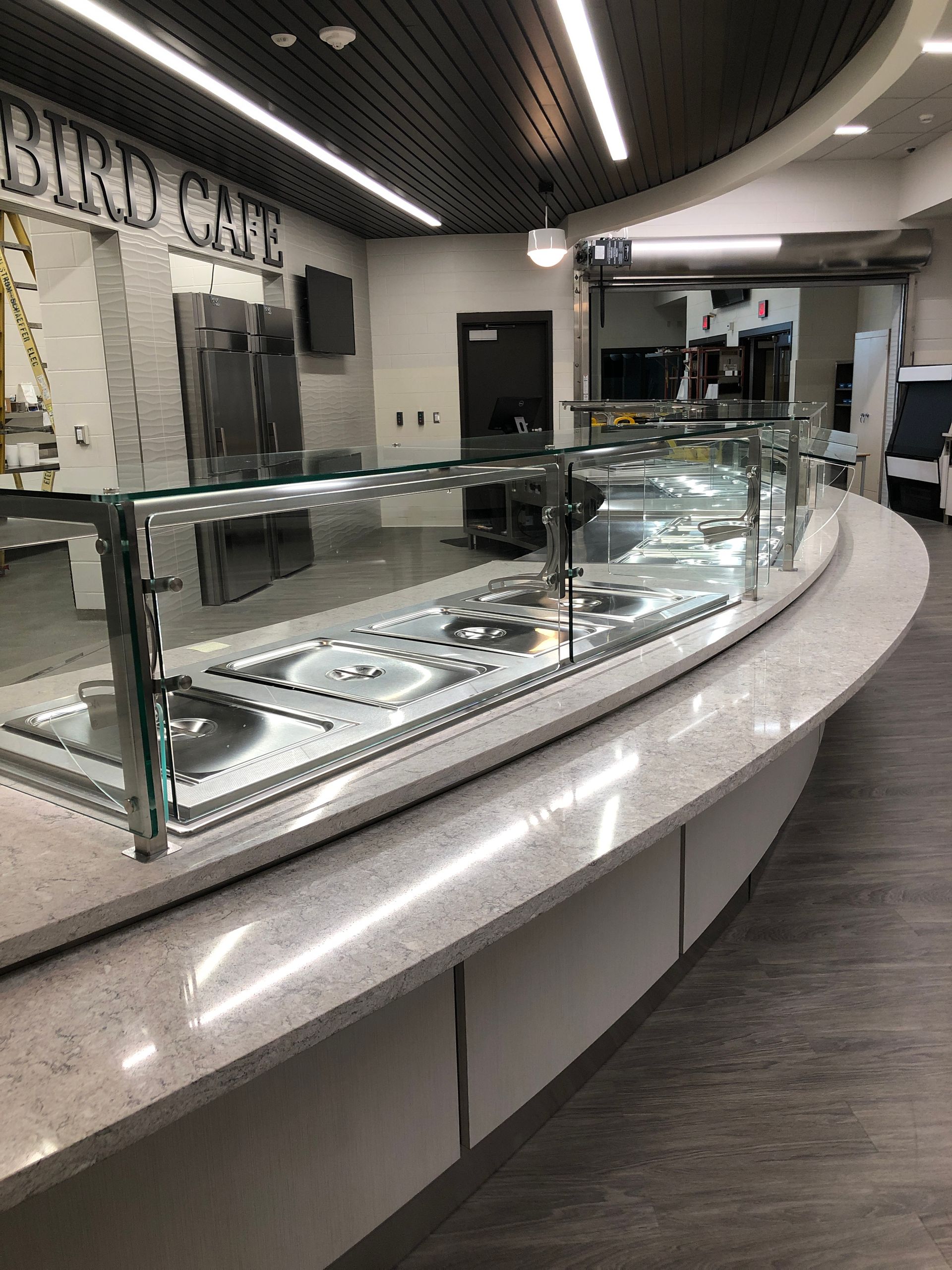 A long curved cafeteria food service counter with a glass sneeze guard and metal serving wells inside a cafe.