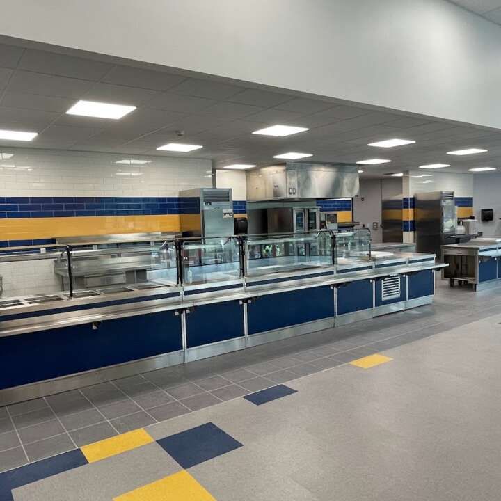 A modern school cafeteria serving area with long stainless-steel counters, blue and yellow tiling, and overhead lighting.