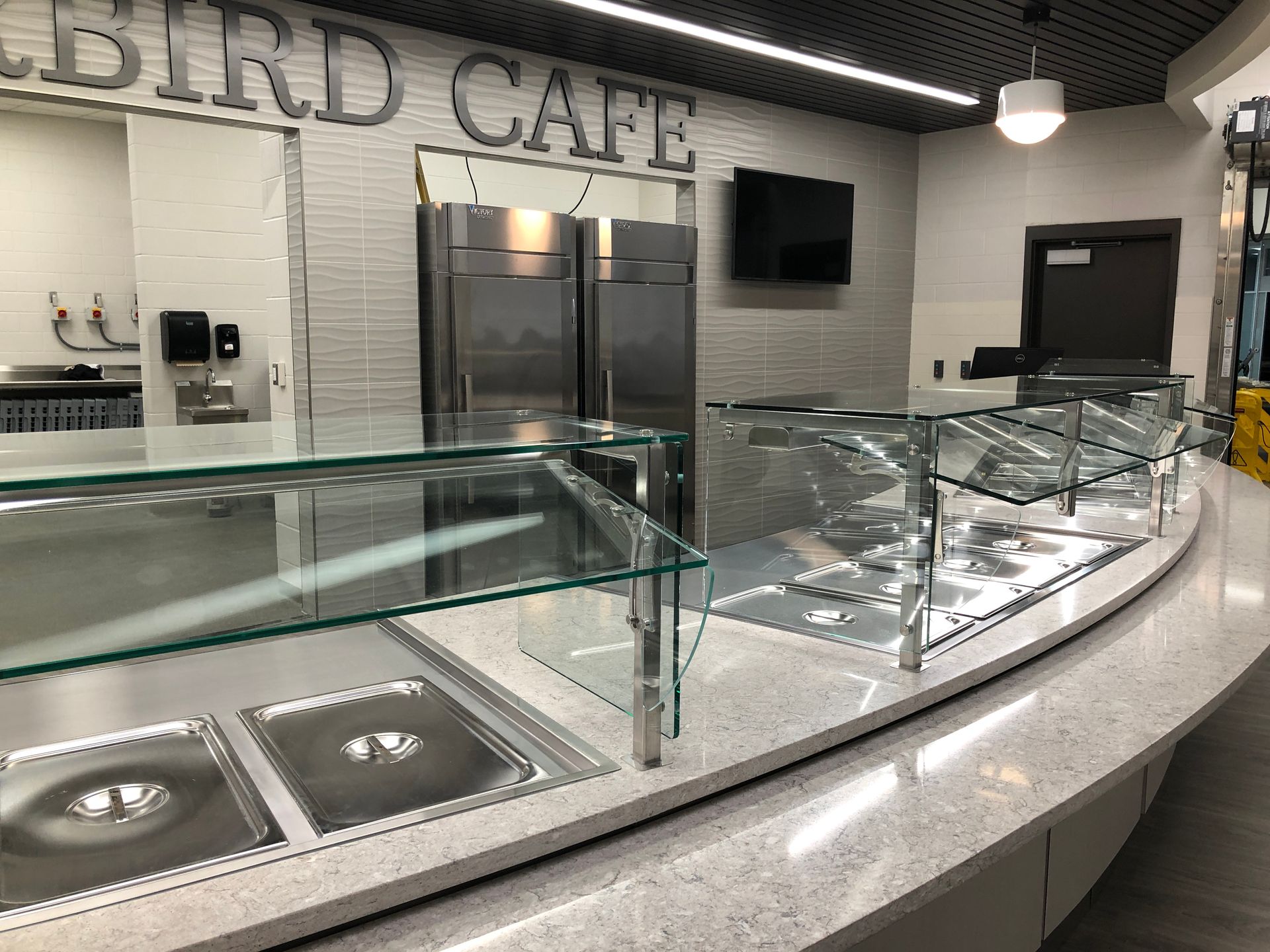 A modern cafeteria service counter with a quartz top, glass sneeze guards, stainless steel food wells, and industrial fridges.
