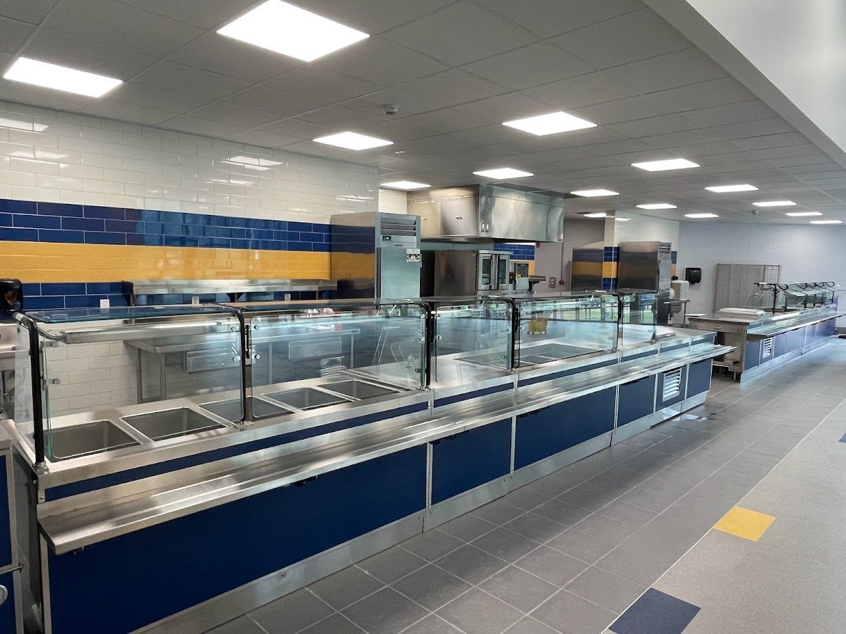 A long, empty cafeteria serving line with stainless steel food wells, glass sneeze guards, and a yellow-and-blue tiled wall.