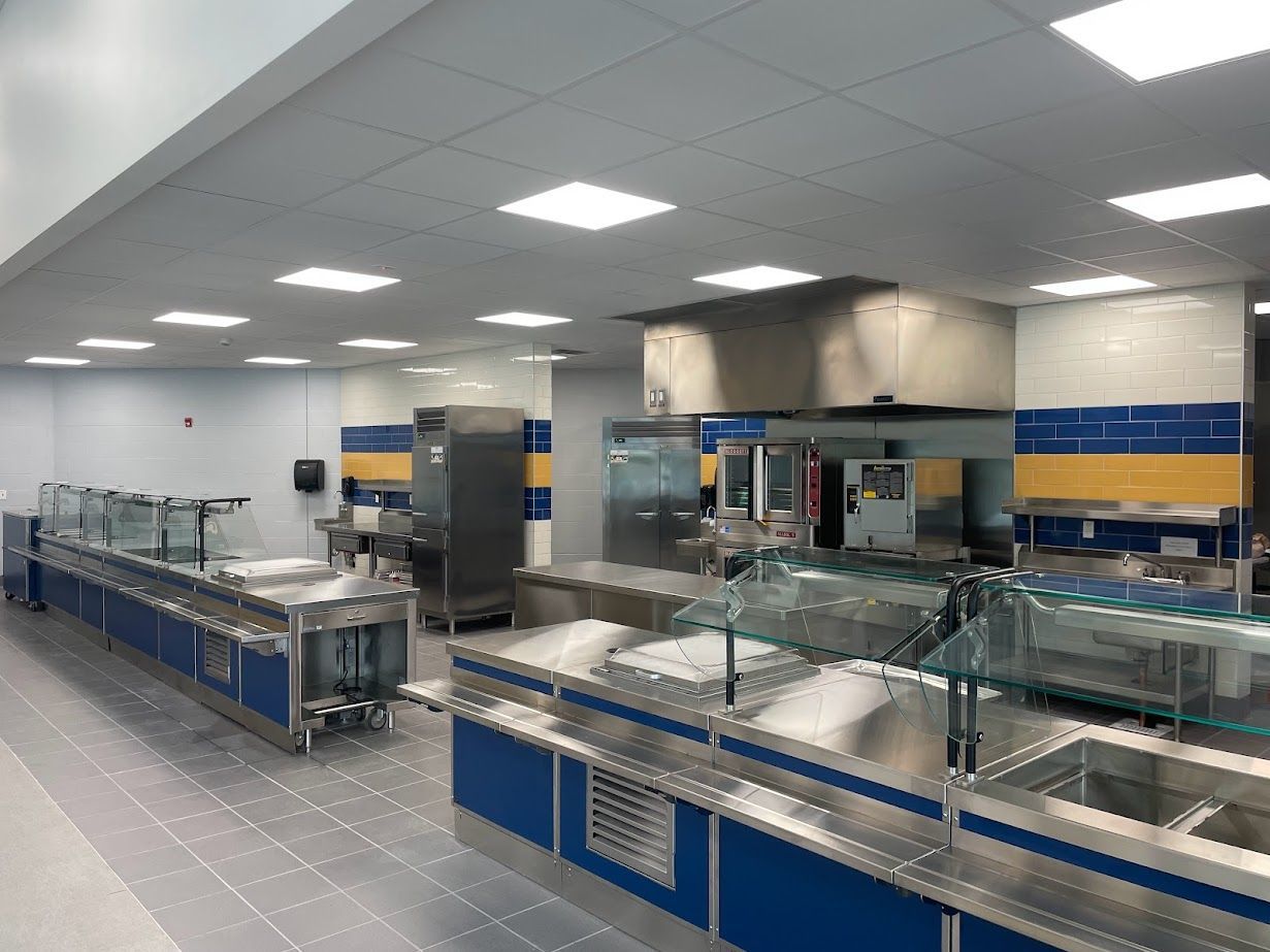 A modern cafeteria serving area with stainless steel food counters, a large exhaust hood, and blue and yellow tiled walls.
