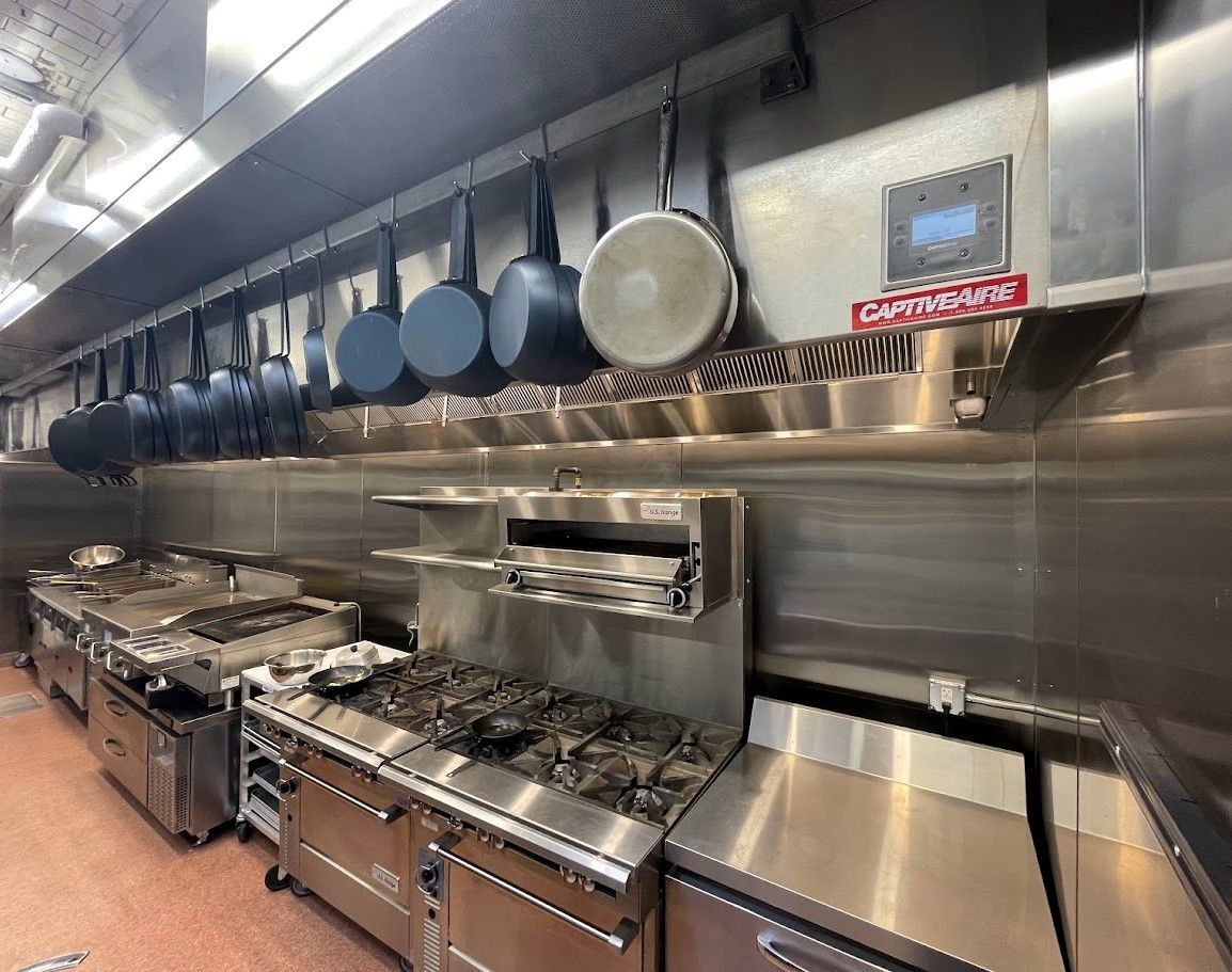 A professional stainless steel kitchen with hanging pots and pans above a row of commercial gas stoves and workspaces.