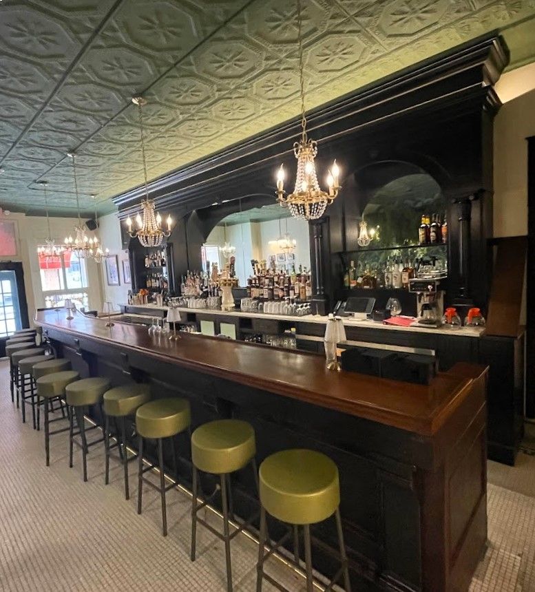 An ornate dark wood bar with olive green round stools, a vintage patterned ceiling, and crystal chandeliers.