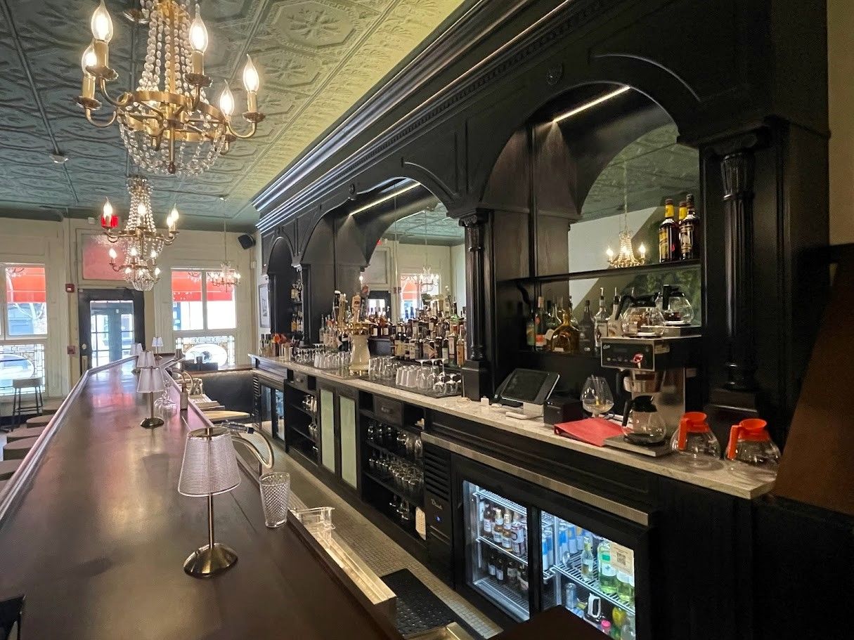 An ornate bar with dark wood cabinetry, a marble countertop, vintage crystal chandeliers, and a patterned teal ceiling.