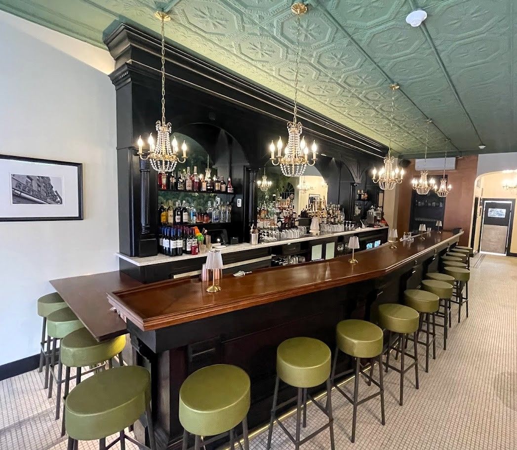 An elegant bar with a polished wood counter, green stools, black shelving, crystal chandeliers, and a patterned ceiling.