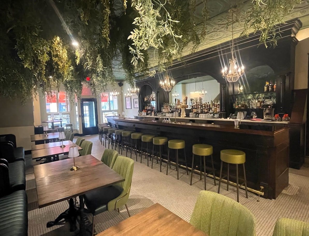 A bar and dining area with dark wooden counters, green bar stools, hanging ceiling foliage, and warm, lit chandeliers.