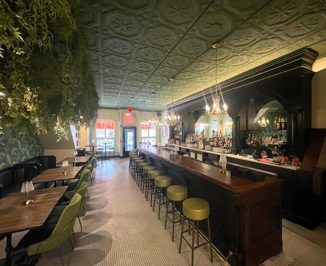 A stylish restaurant interior featuring a dark wood bar with stools, green-patterned ceiling, and cozy dining tables.