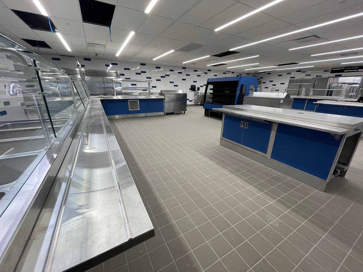A modern, clean cafeteria space with stainless steel serving counters, blue base cabinets, and gray tile flooring.