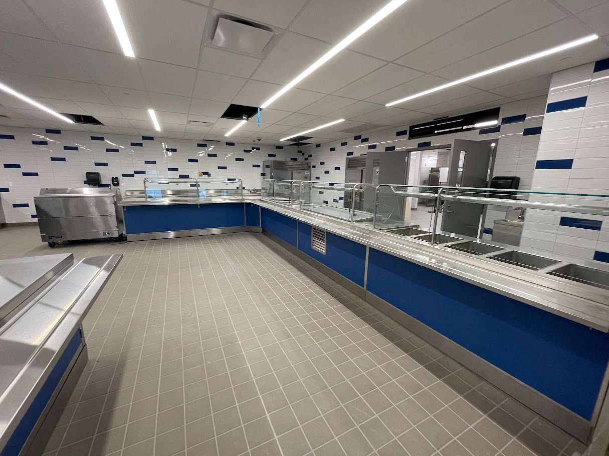 A modern cafeteria service line featuring a long blue counter, stainless steel food wells, and blue-and-white wall tiles.