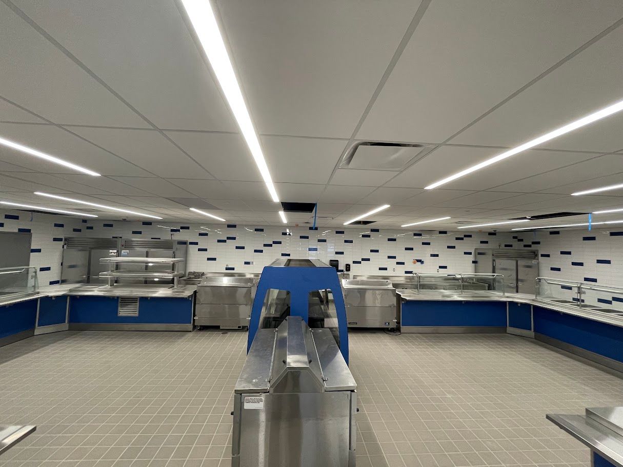 Empty cafeteria serving area with stainless steel counters, blue accents, and a tile wall with scattered black rectangles.