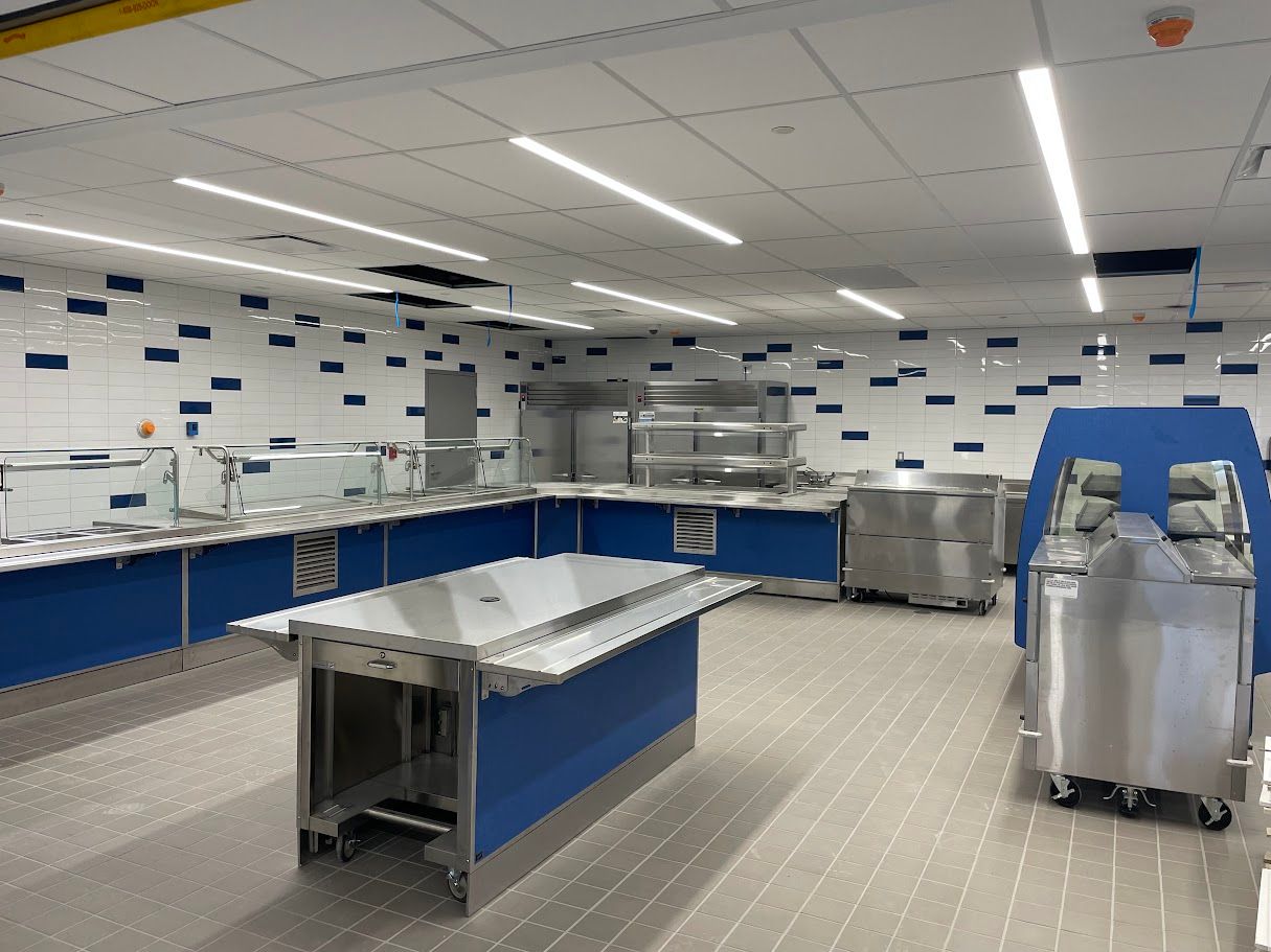 A modern, empty school or commercial cafeteria with blue counters, stainless steel equipment, and white tiled walls.