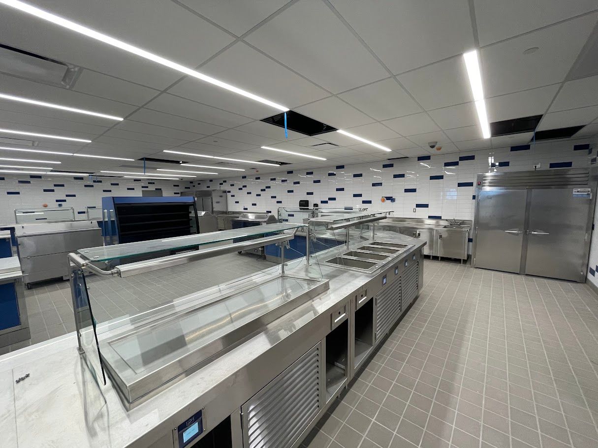 A brightly lit, modern commercial kitchen with stainless steel prep counters, serving stations, and tiled walls.