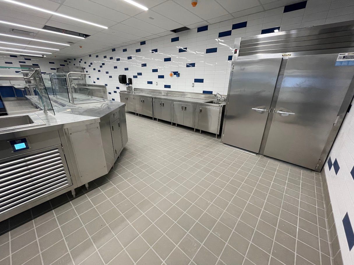 Commercial kitchen with stainless steel equipment, tiled walls with blue accents, and grey floor tiles.