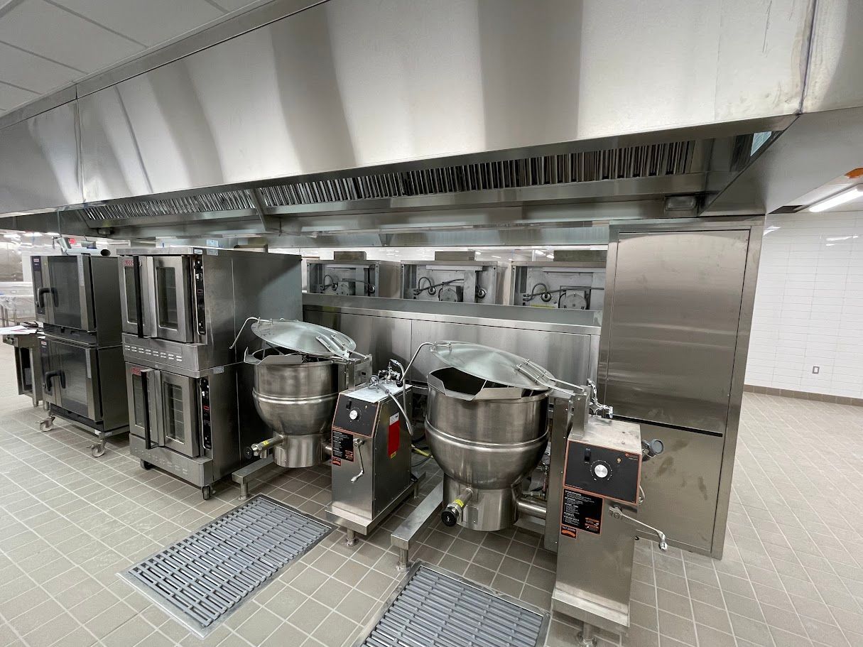 A commercial kitchen line featuring stainless steel ovens and two large, floor-mounted steam kettles under a vent hood.