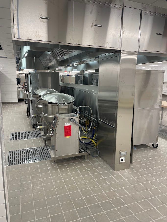 A commercial kitchen features stainless steel industrial cooking pots and vent hoods under overhead cabinetry.