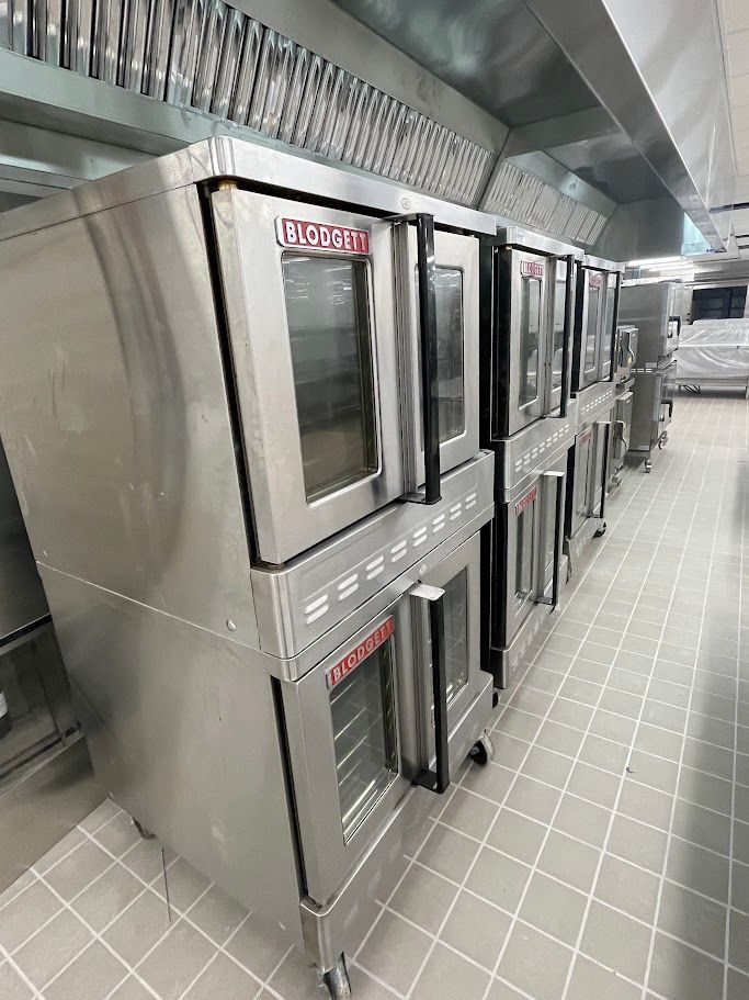 A row of stainless steel commercial double-stack convection ovens in a kitchen with tiled floors.