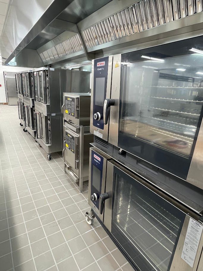 A row of stainless steel commercial combi ovens under a ventilation hood in a professional kitchen with tiled flooring.