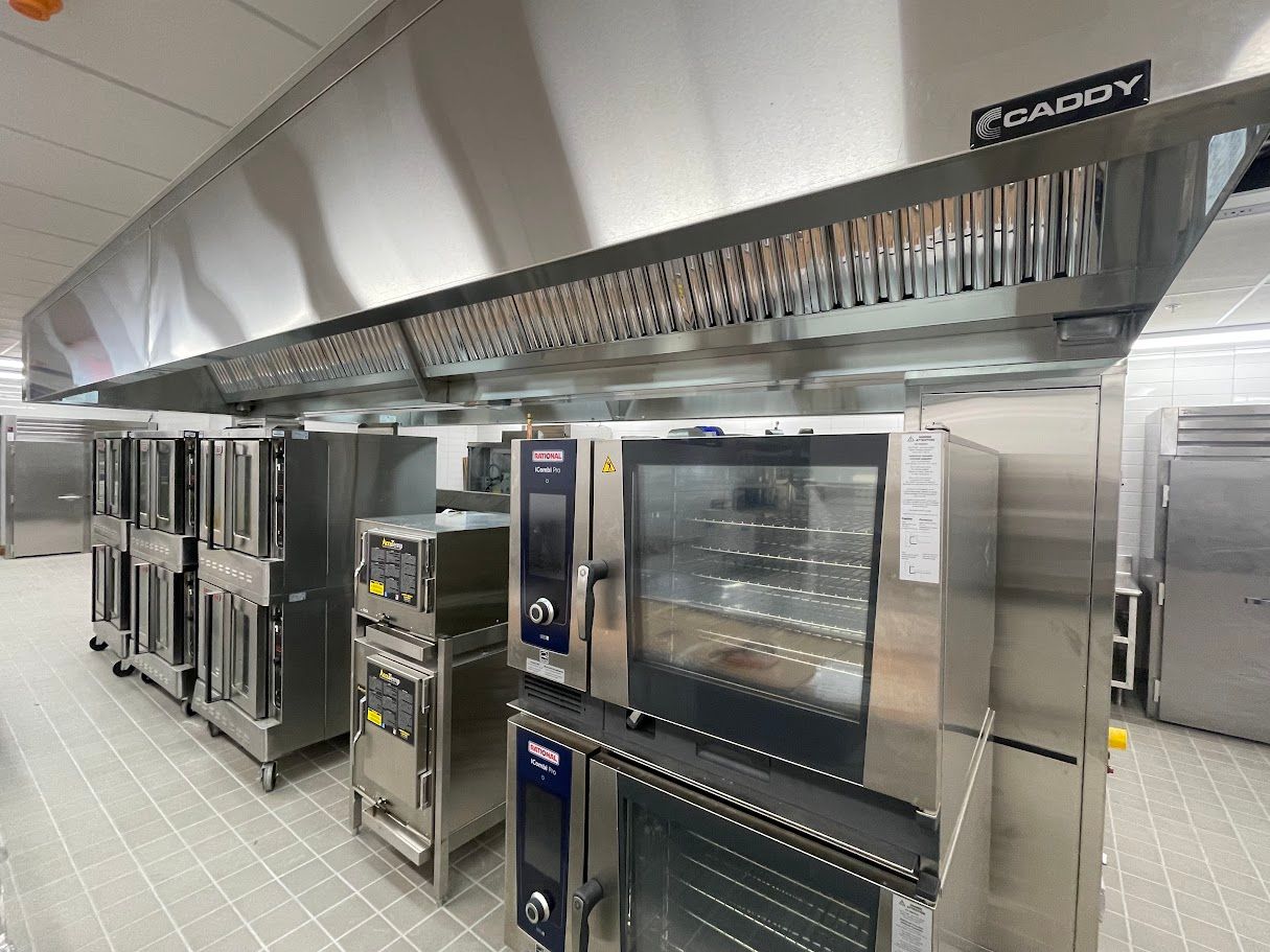 A commercial kitchen line with rows of stainless-steel ovens under a Caddy ventilation hood.