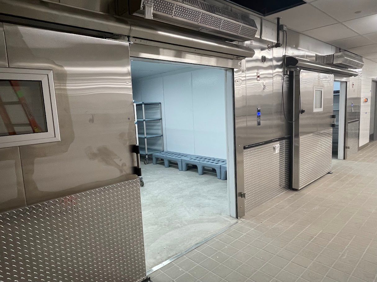 A commercial walk-in cooler or freezer with stainless steel walls and two sliding doors, one open showing interior shelving.