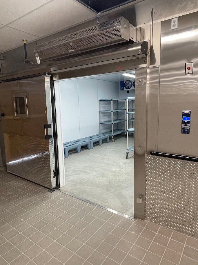 A brightly lit industrial walk-in freezer with metal shelving, floor pallets, and a sliding door.