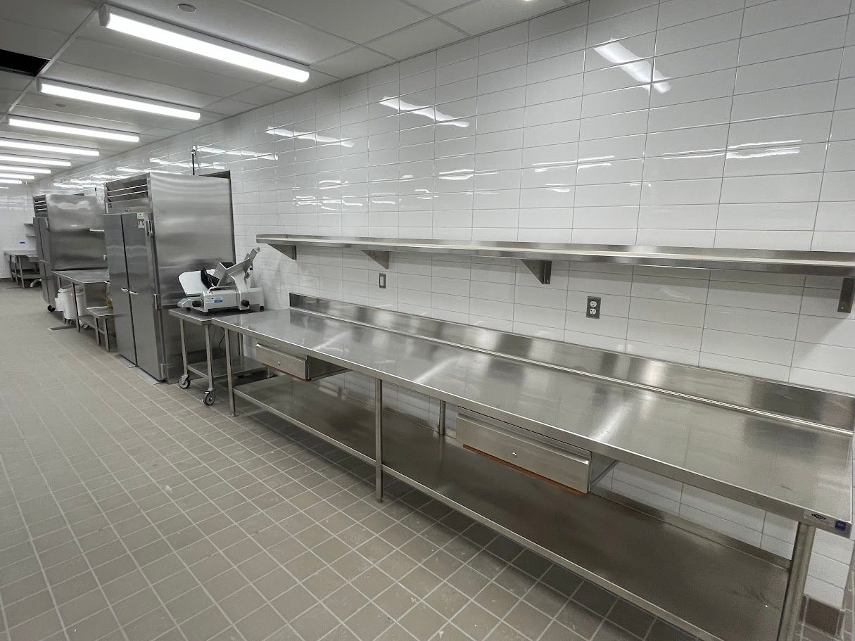 A commercial kitchen with stainless steel tables, prep equipment, and a white tiled wall.