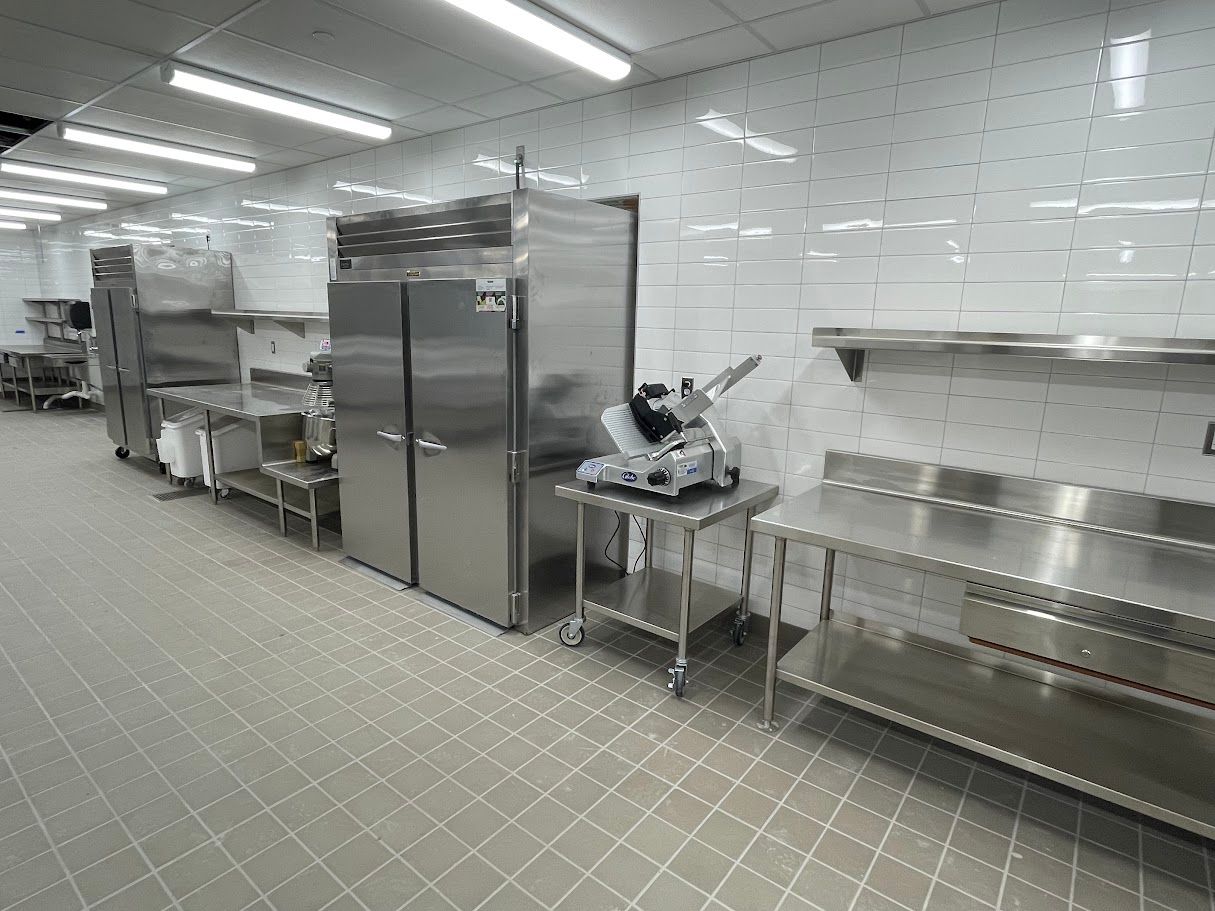 Commercial kitchen with stainless steel refrigerators, food prep tables, and a slicer on a tiled floor and walls.