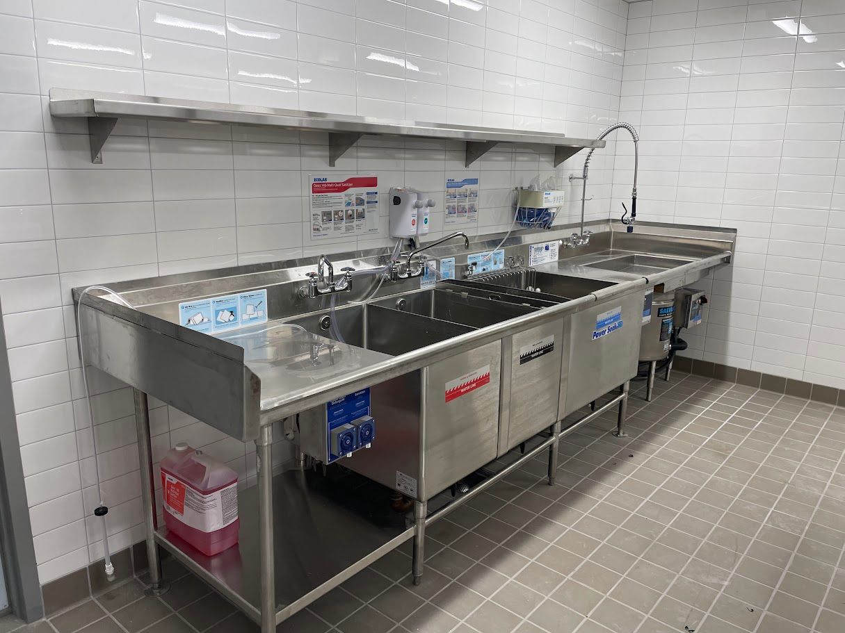 A commercial stainless steel, three-compartment sink setup with a faucet, dispensers, and a lower shelf against tiled walls.