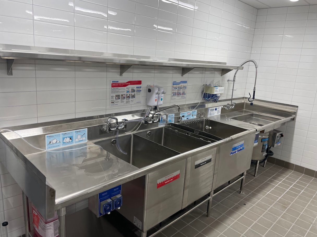 A stainless steel multi-compartment sink in a commercial kitchen with tiled walls and an overhead shelf.