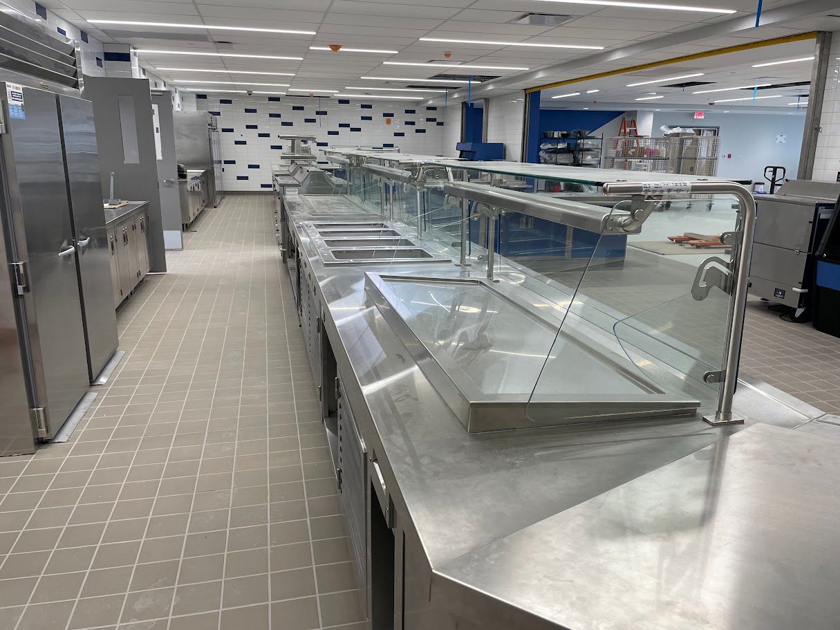 A long, empty commercial kitchen serving line with stainless steel counters and glass sneeze guards.