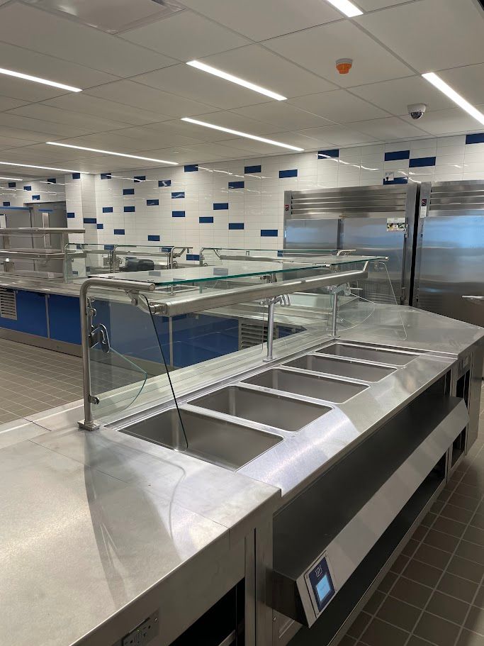 A stainless steel food service buffet counter with glass partitions in a clean, modern cafeteria with tiled walls.