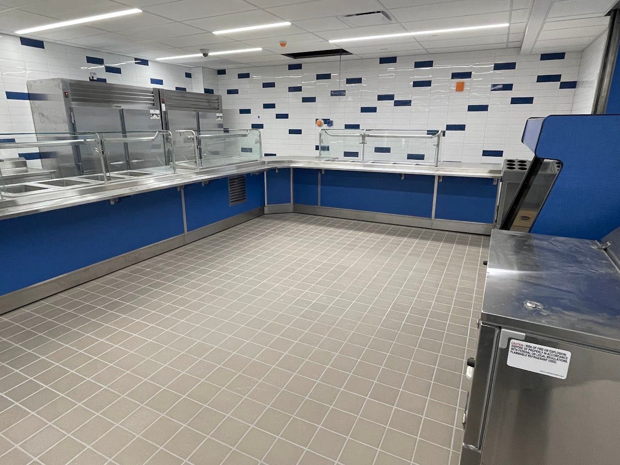 A modern cafeteria serving area with stainless steel food counters, a blue base, and a white tiled wall with blue accents.