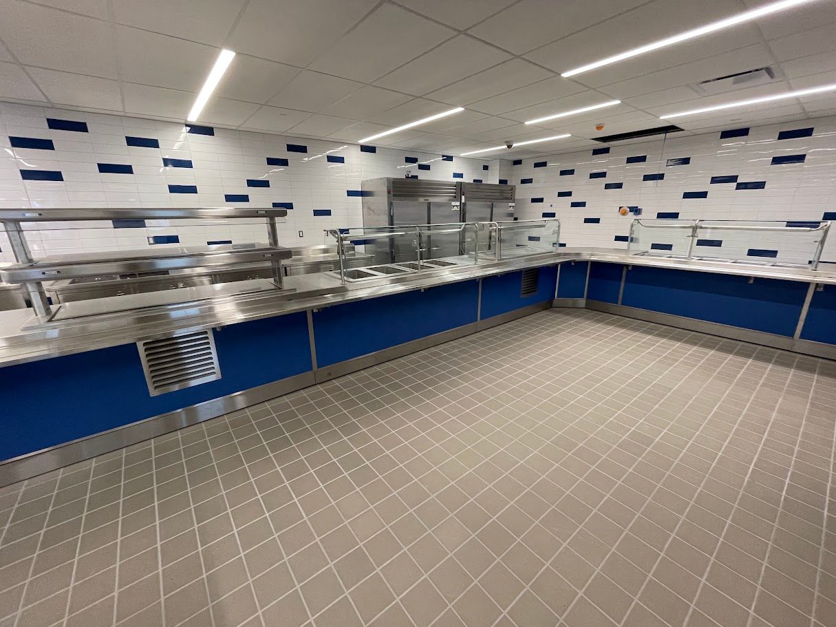 Empty cafeteria service counter with stainless steel equipment, tiled walls with blue accents, and grey floor tiles.