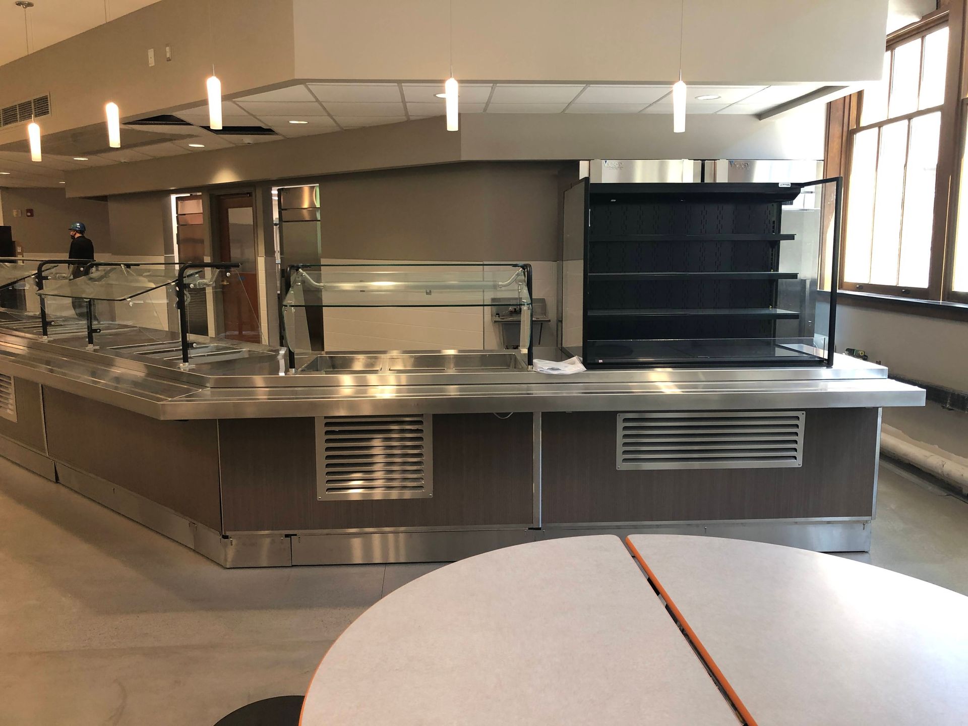An empty cafeteria serving line with a stainless steel counter and a black refrigerated display case in a dining room.