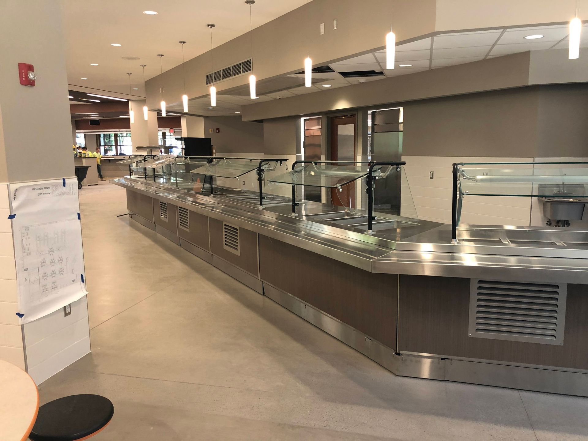 A long, stainless-steel food service counter with glass sneeze guards in a modern, brightly lit cafeteria dining area.