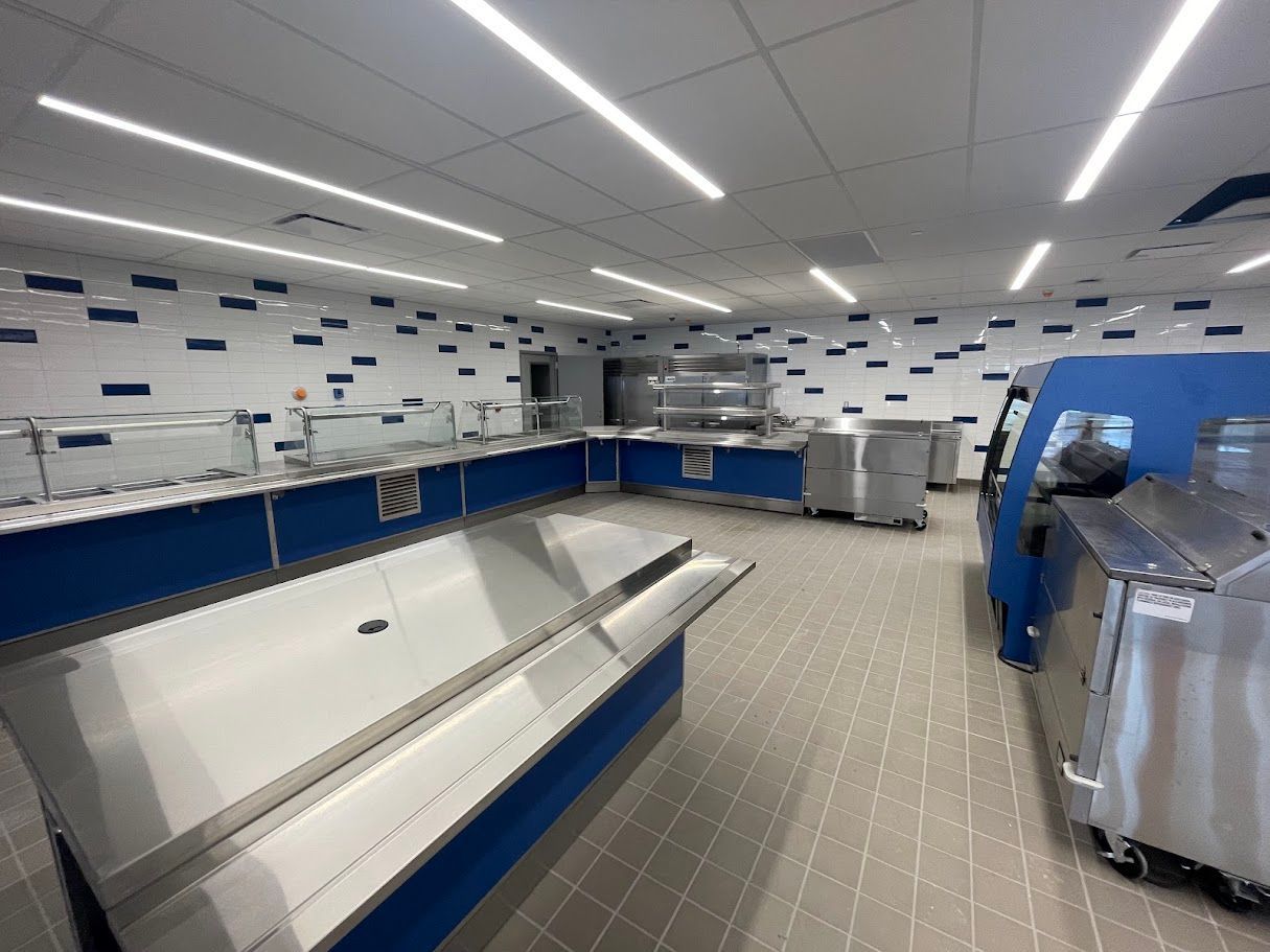 A modern cafeteria serving area with stainless steel food counters, blue accents, and tiled flooring under bright lights.
