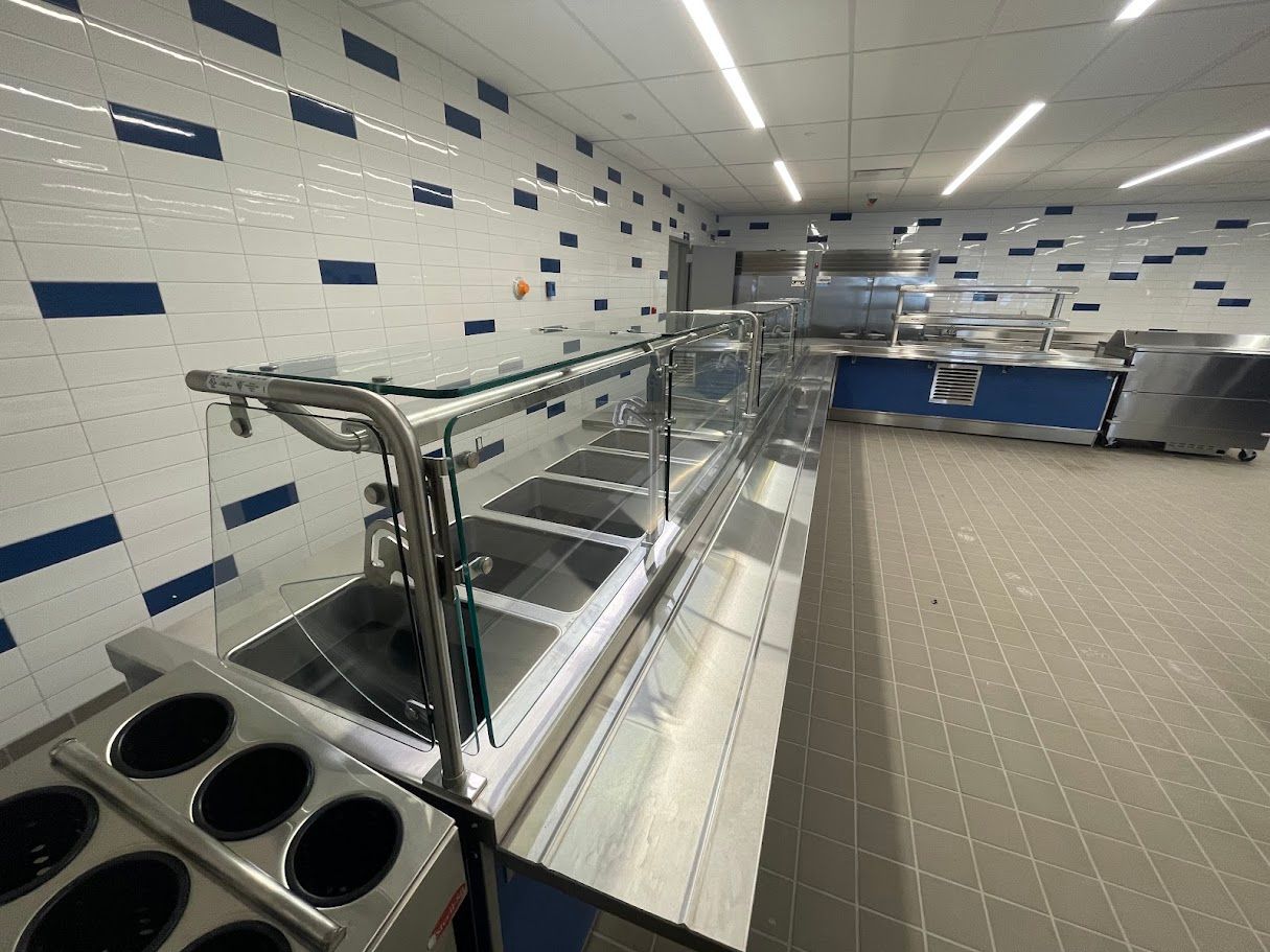 A commercial food service counter with glass sneeze guards, tiled walls with blue accents, and metallic serving stations.