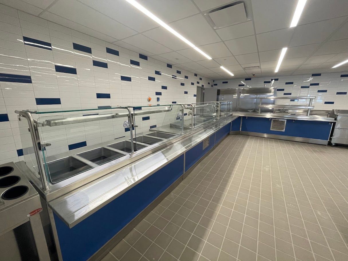 Empty cafeteria serving line with stainless steel food wells, glass sneeze guards, and blue accents on a tiled floor.