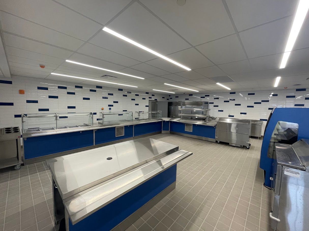 A brightly lit, modern cafeteria serving line with stainless steel counters and blue cabinetry against tiled walls.