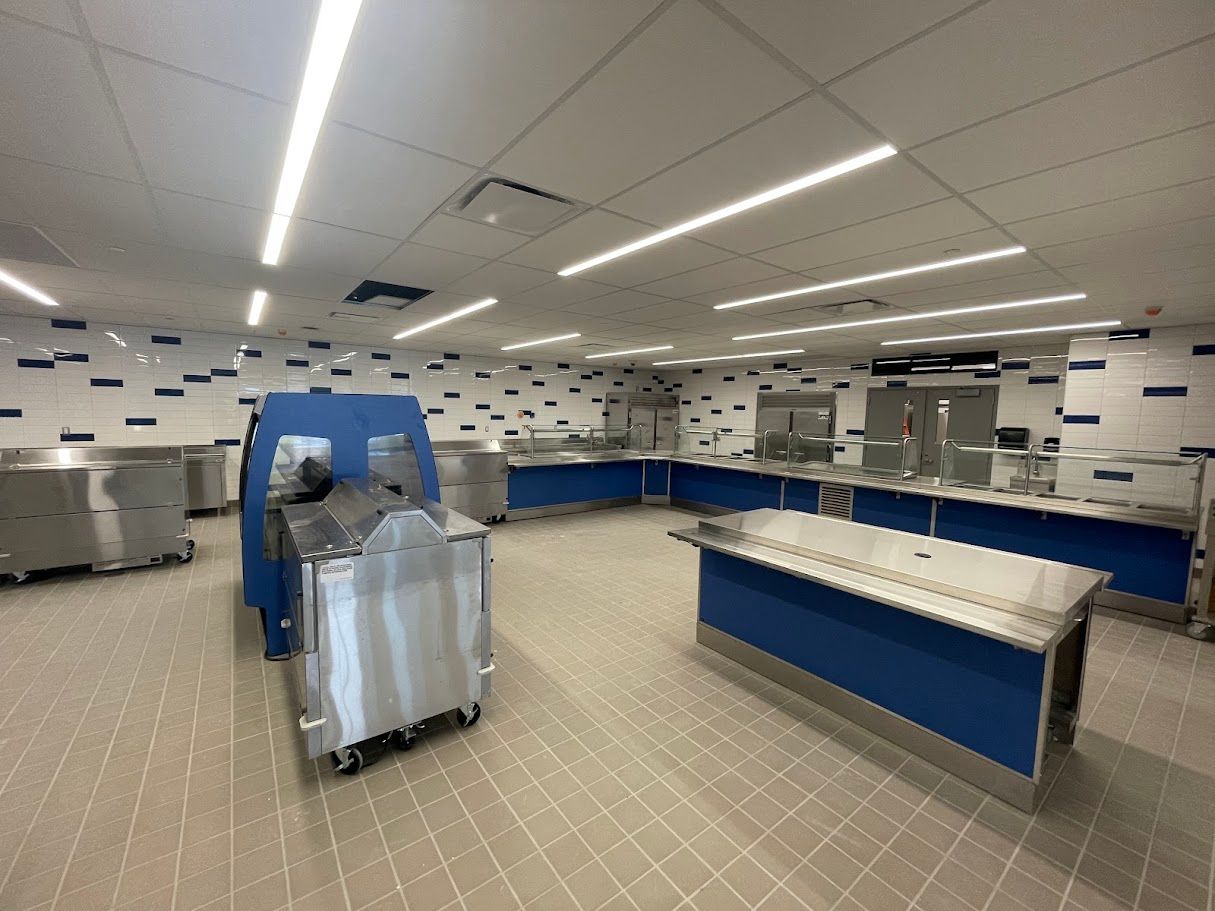 A commercial kitchen features stainless steel prep tables, blue serving counters, and tiled walls with blue accents.
