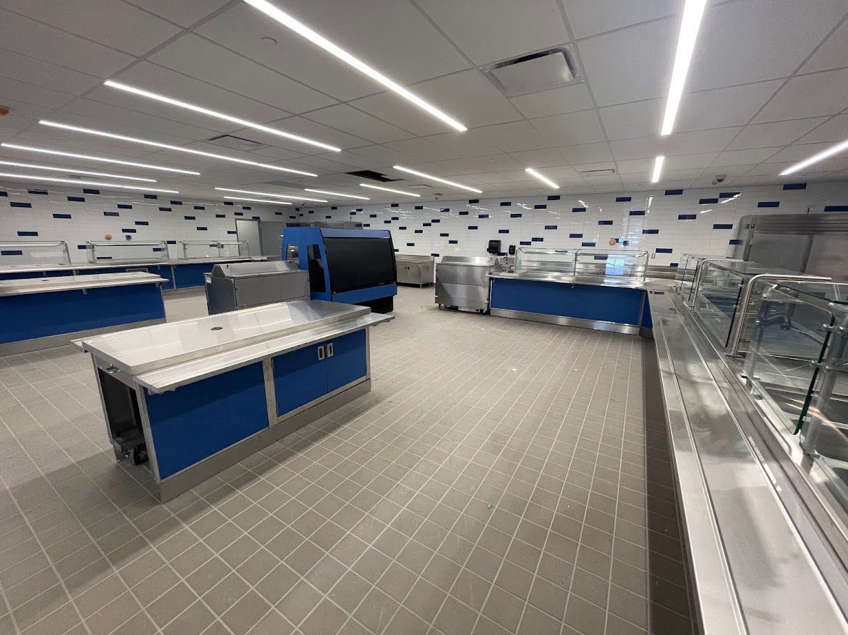 A modern, clean cafeteria space featuring blue serving stations and tables on a tiled floor under bright ceiling lights.