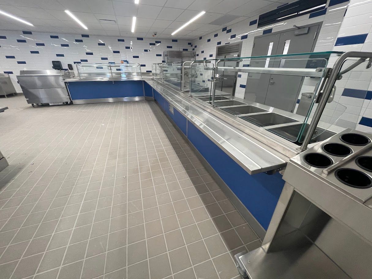 Empty school cafeteria serving line with blue cabinetry, stainless steel counters, and white tiled walls.