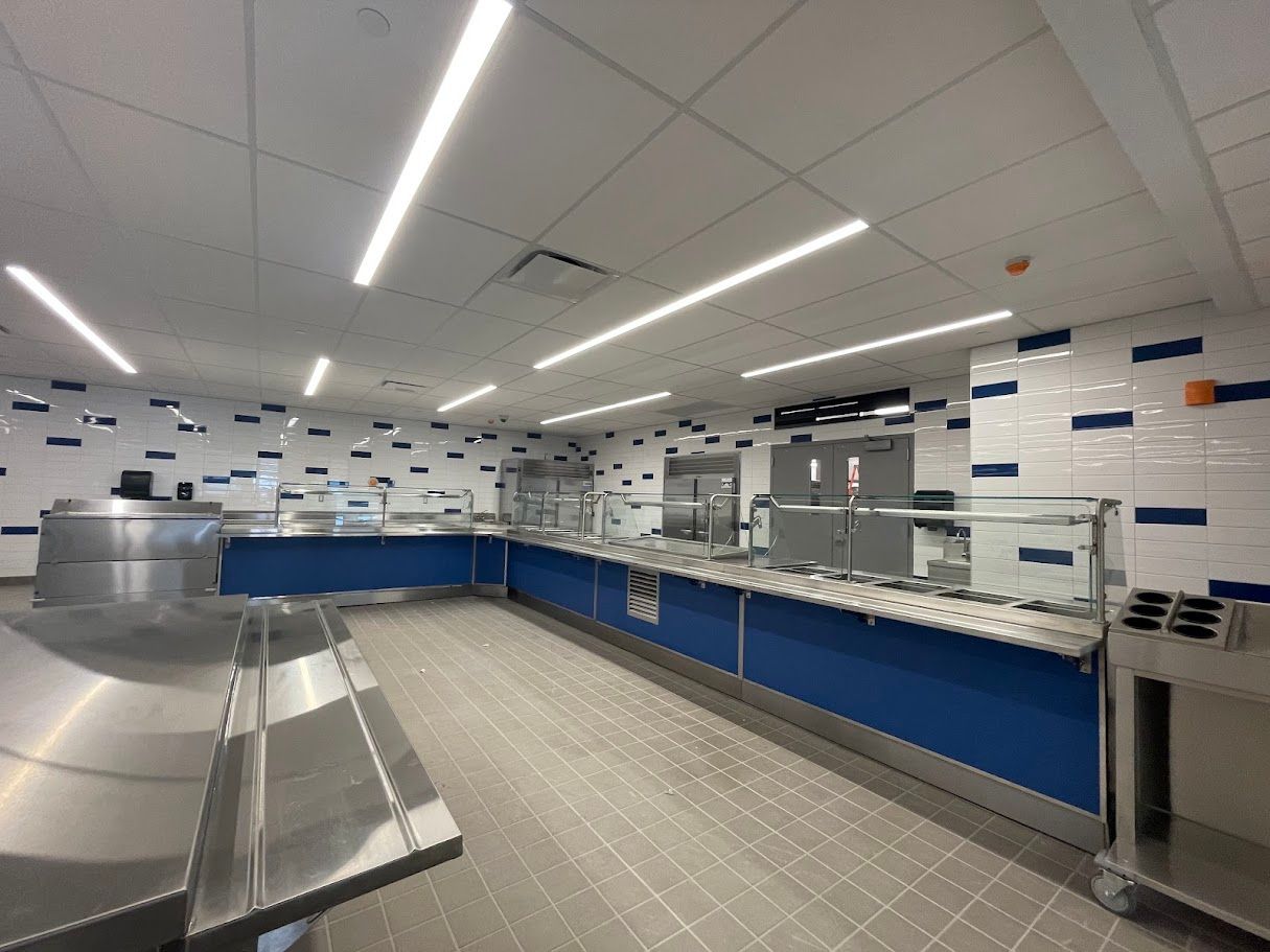 A spacious commercial cafeteria service line with blue counters, stainless steel food stations, and white tiled walls.